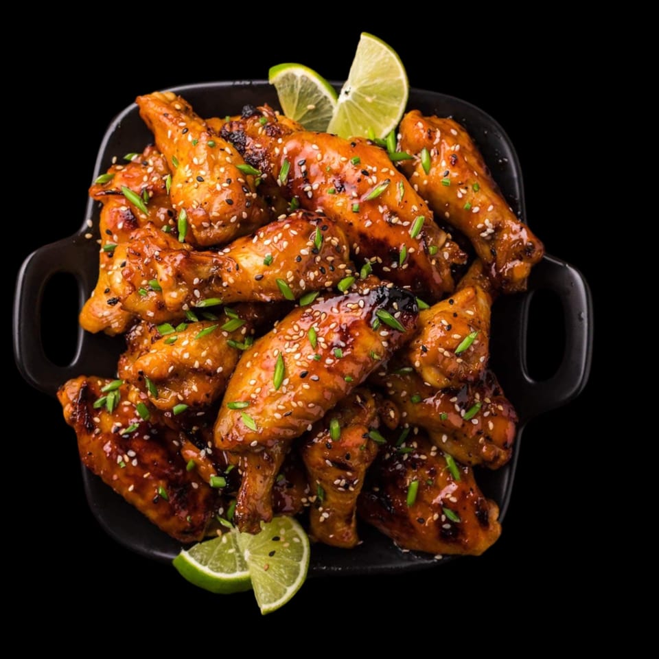 Glazed chicken wings with sesame seeds, chives, and lime wedges in a black bowl.