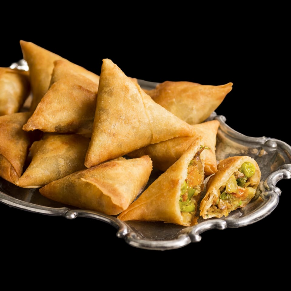 Golden-brown samosas, one cut open to reveal a savory filling, on a decorative silver platter.